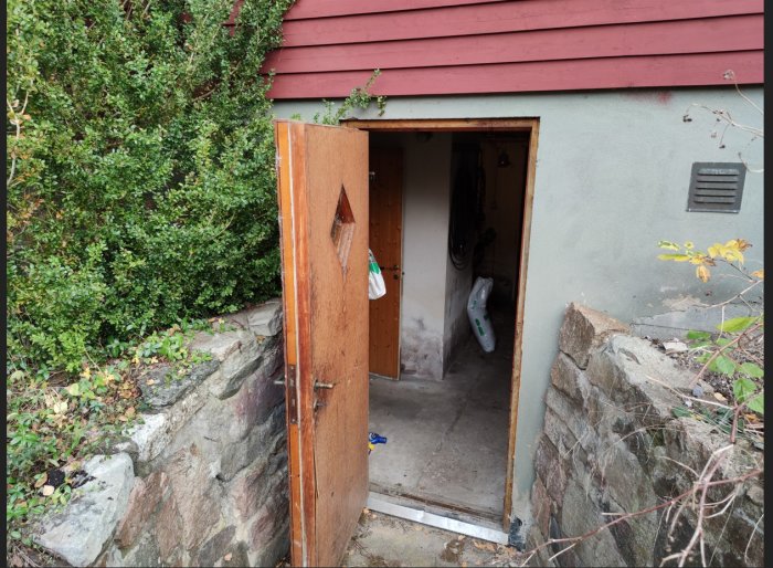 Öppen källardörr till ett rött hus, omgivet av grönt buskage och en stenmur, vilket visar tröskeln och delvis interiören.