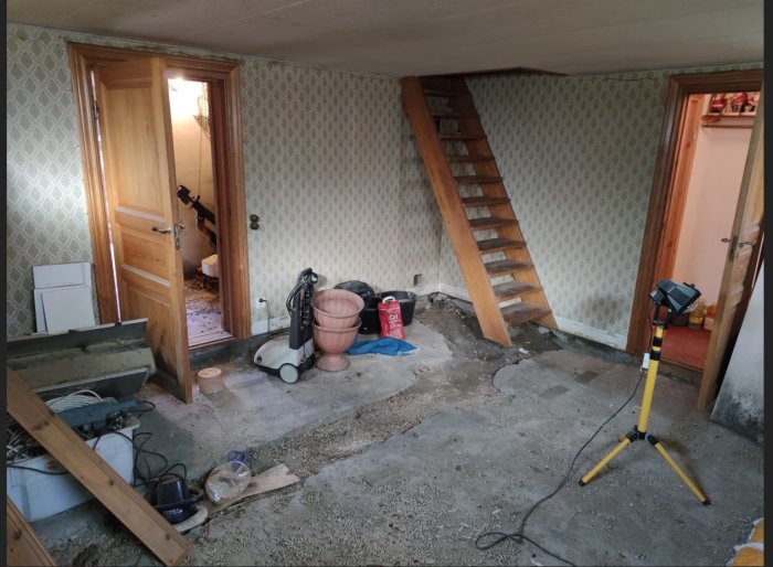 Orenoverad källare med utbrytet golv, byggavfall, verktyg och en trappa i ett fritidshus under renovering.