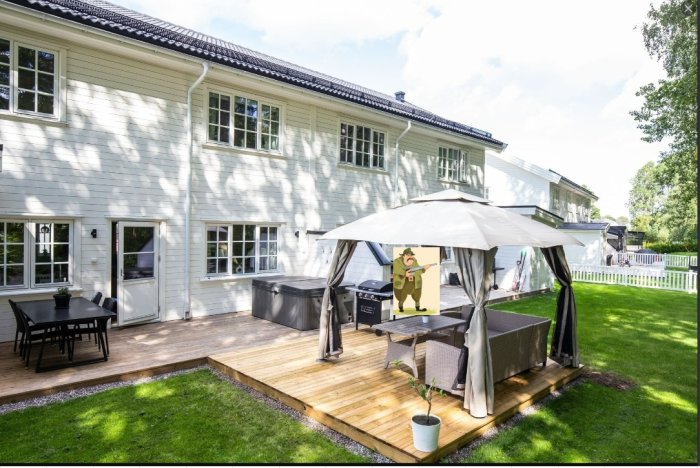 Vit tvåvåningsvilla med terrass, utemöbler och grill under paviljong samt en tecknad figur.