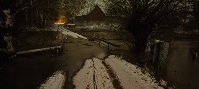 Översvämmad körbana med snö och översvämmande ån vid gryning, med träd och byggnad i bakgrunden.