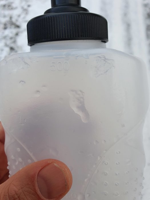 Hand håller en genomskinlig vattenflaska med kondens utomhus i vintermiljö.