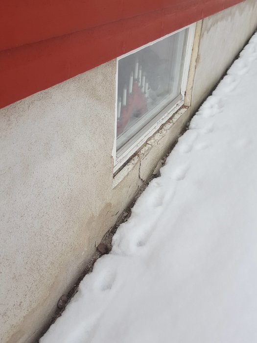Skadad fasadputs nära ett fönster med synlig sprickbildning och avskalning ovanför snötäckt mark.