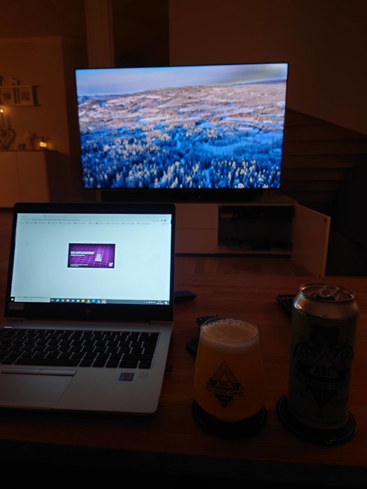 En hemarbetsplats med en bärbar dator, ett ölglas och en burk från Apex Brewery, framför en TV som visar en naturbild.