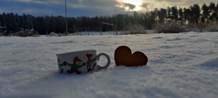 Julmugg och pepparkaka i hjärtform på ett snötäckt landskap med solnedgång i bakgrunden.