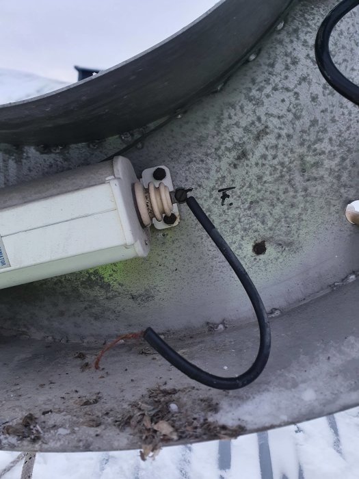 Elektrisk kabel går genom en vit plastklämma fäst på en metallränna med smuts och lövavfall.