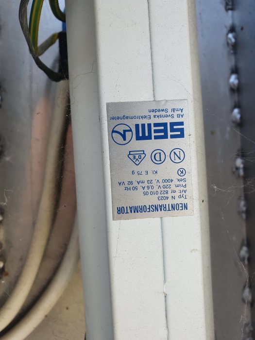 Etikett med specifikationer på en elektrisk enhet, monterad upp och ner, med synliga kablar och spindelväv.