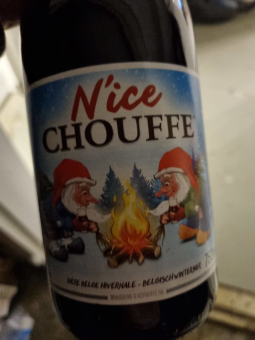 Flaska N'ice Chouffe julöl med etikett visande två tomtenissar som värmer sig vid en eld under snöfall.