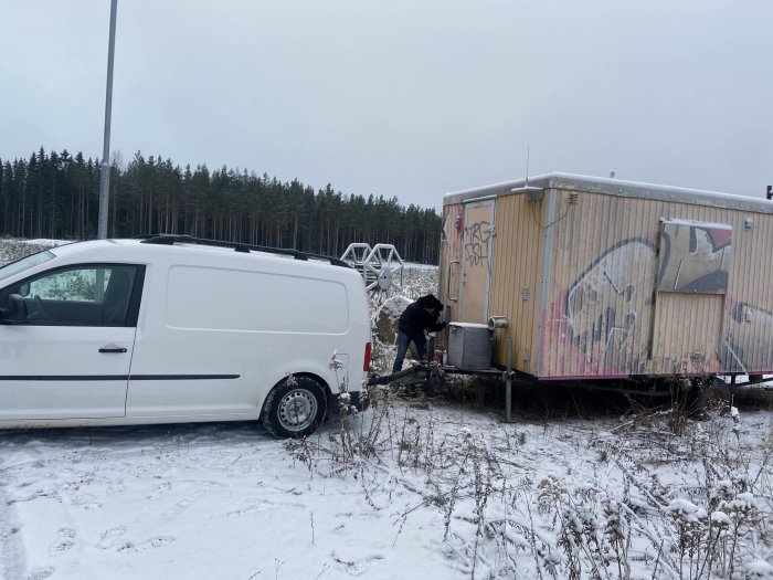 Vit skåpbil kopplad till en sliten släpvagn med graffiti på ett snötäckt fält.