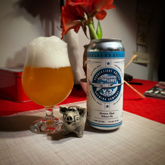 Ölglas fyllt med skummande öl bredvid burk och dekorativ grisfigur på rödvit duk.