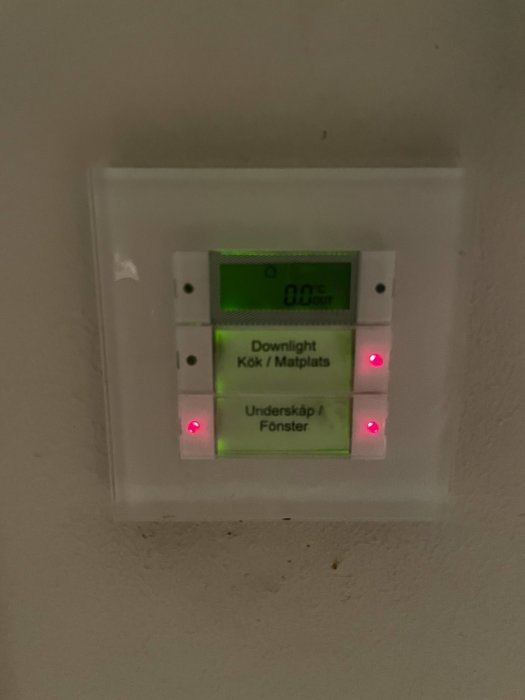 Väggmonterad KNX strömställare/termostat med textetiketter "Downlight Kök/Matplats" och "Underskåp/Fönster" tända röda indikatorlampor.