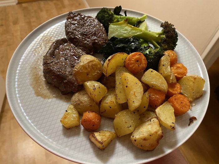 Två stycken grillade rostbiffar med ugnsrostade potatisar, morötter, broccoli och rödlök på en tallrik.