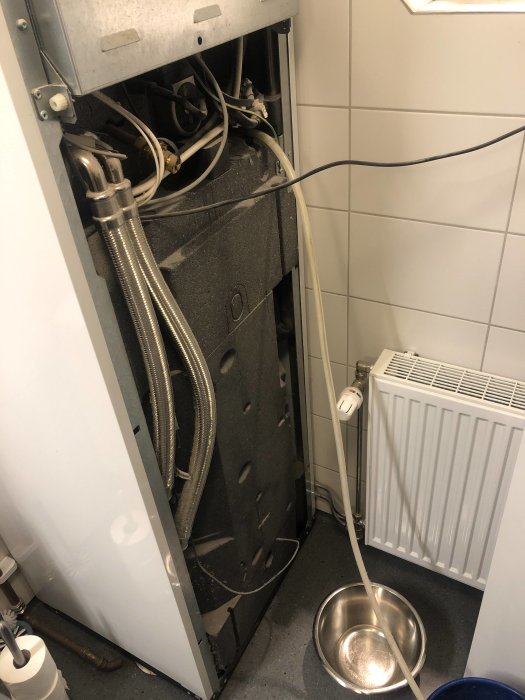 Avmonterad diskmaskin med öppen baksida som avslöjar rör och ledningar, placerad i ett vitt kaklat hörn intill en radiator.