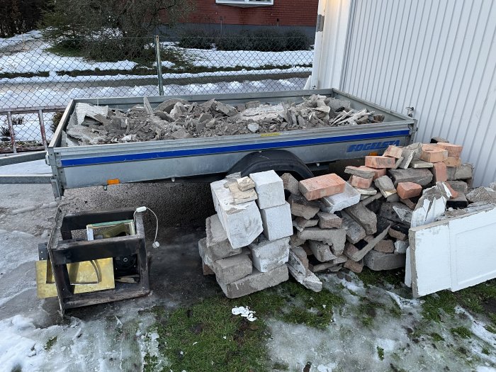 En släpvagn fylld med rivningsavfall och en hög med tegelstenar bredvid, med en gammal kronspis på marken framför.