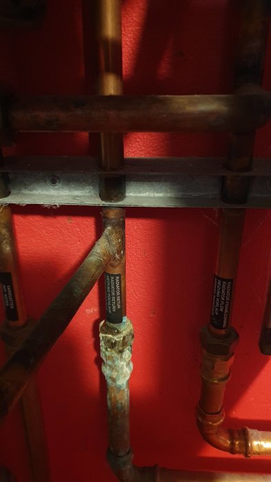 Kopparrör med etiketter och en synlig läcka, mot en röd vägg.