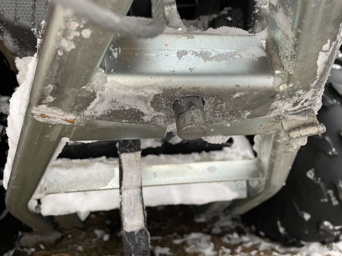 Del av fyrhjulingsplog med saknad sprint och synliga skador på fästet, täckt av snö.
