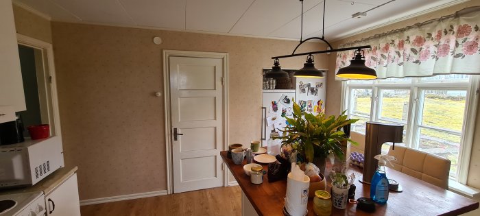 Kök före renovering med daterad tapet och köksbord fyllt med hushållssaker.