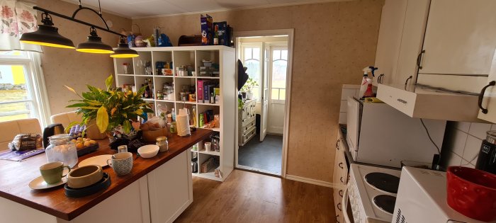 Gammalt kök före renovering med träarbetsbänk, öppna hyllor och ljusa inslag, innan rivning av vägg.