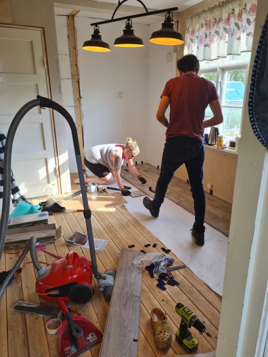 Två personer installerar nytt golv i ett rum under renovering, omgivna av verktyg och byggmaterial.