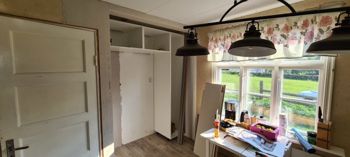 Ett kök under renovering med omonterade skåpstommar och verktyg på arbetsbänken, med ett fönster som vetter mot en gård.