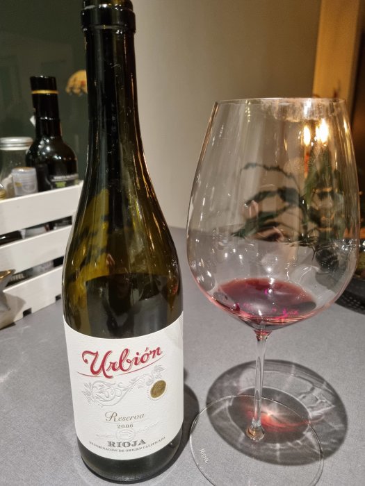 Flaska Rioja-vin och ett halvfyllt glas på bord, diffust synlig flaska i bakgrunden.