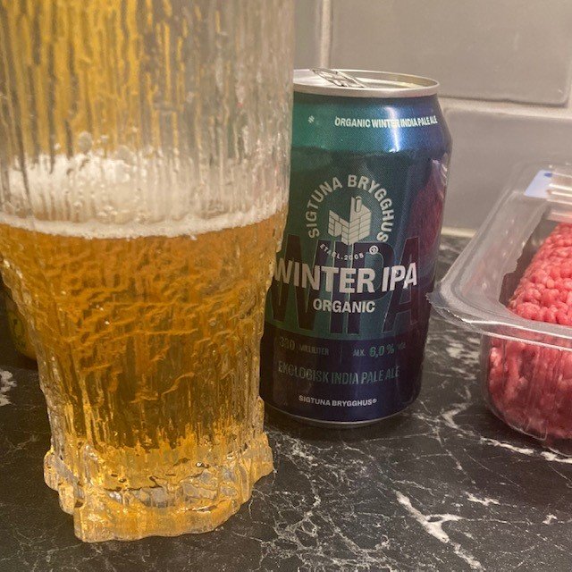 Halvtomt glas med gyllene öl framför en Sigtuna Winter IPA-burk och en låda med hallon.