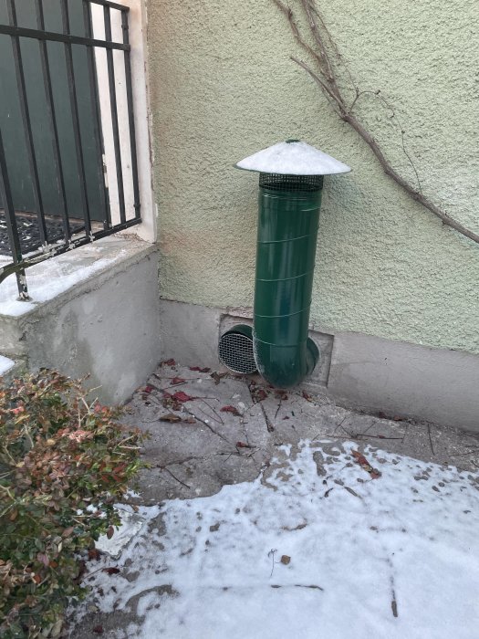 En grön luftventil utifrån ett hus, i ett hörn nära marken med snö och ett skyddsgaller på marken.