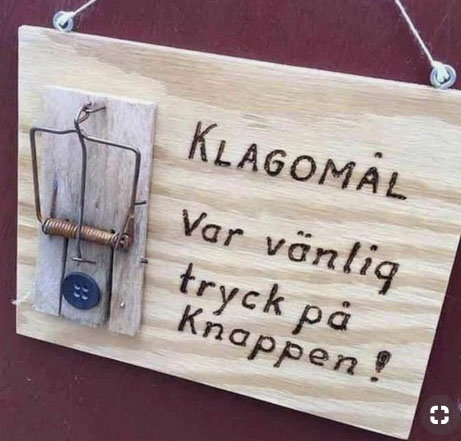 Träskylt med texten "KLAGOMÅL Var vänlig tryck på knappen" och en ironiskt menad "knapp" av en musfälla.