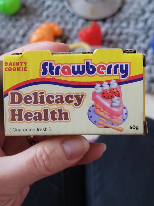 En leksakskartong för "Strawberry Delicacy Health" kex, hållen i en hand med suddig bakgrund.