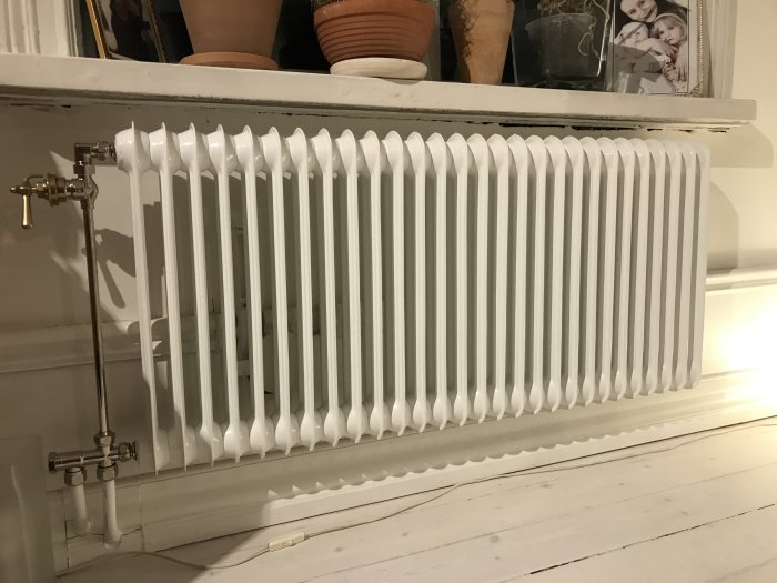 Vit sektionradiator monterad intill en vägg under ett fönster med krukväxter ovanför.