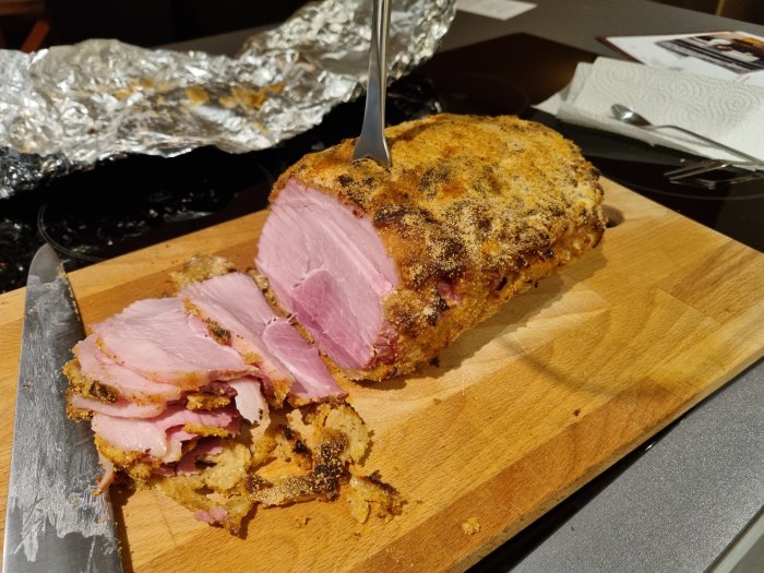 Julskinka skuren på skärbräda med kniv och aluminiumfolie i bakgrunden.