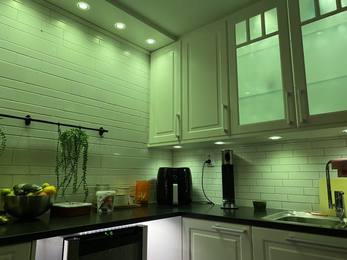Kök med grönt ljussken från spottar i taket, vita skåp och tegelvägg, köksutrustning på bänkskiva.