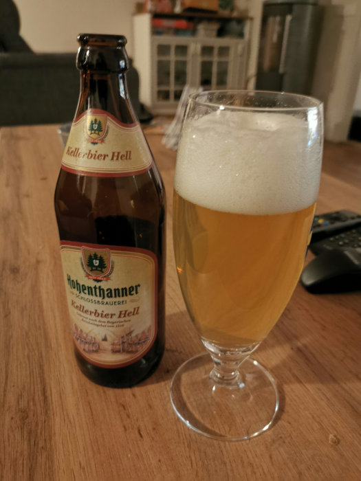Flaska av Hohenthanner Kellerbier Hell bredvid ett glas med öl, på ett bord i ett vardagsrum.
