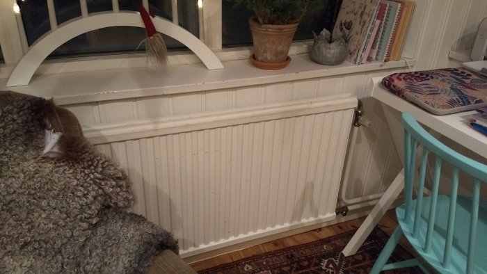 Radiator under fönster i ett äldre hem med inredningsdetaljer och en blå stol i förgrunden.