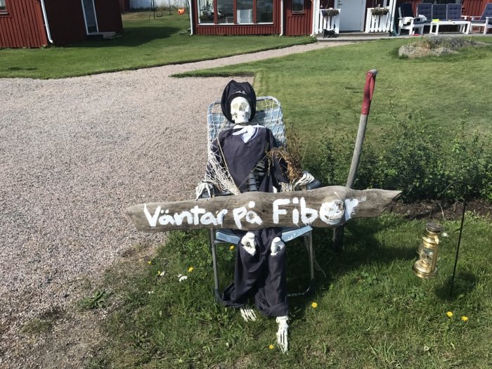 Skämtsam installation av en skelettdocka sittandes i en stol med skylt "Väntar På Fiber".