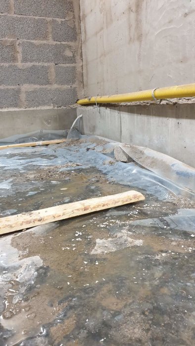 Källargolv med vattensamlingar ovanpå plastduk, träplankor och makadam mot betongväggar.
