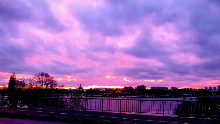 Solnedgång med dramatisk rosa och lila himmel över en stadssilhuett och flod.