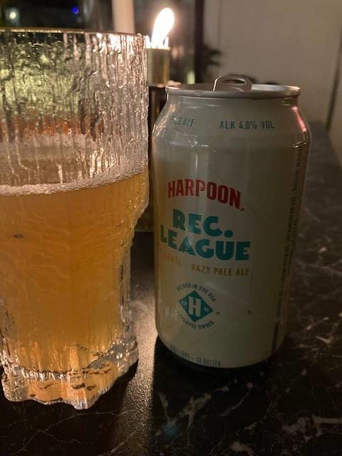 Öppnad burk av Harpoon Rec. League Hazy Pale Ale med ett halvtomt glas öl bredvid, på ett bord.