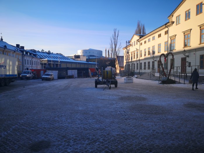 Tomt torg med byggnader och byggmaterial en solig dag, snötäckt mark och en person som går.