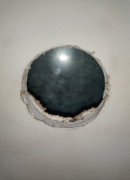 Ett runt hål sågat i ett vitt innertak med ojämna kanter och mörkt utrymme synligt genom öppningen.
