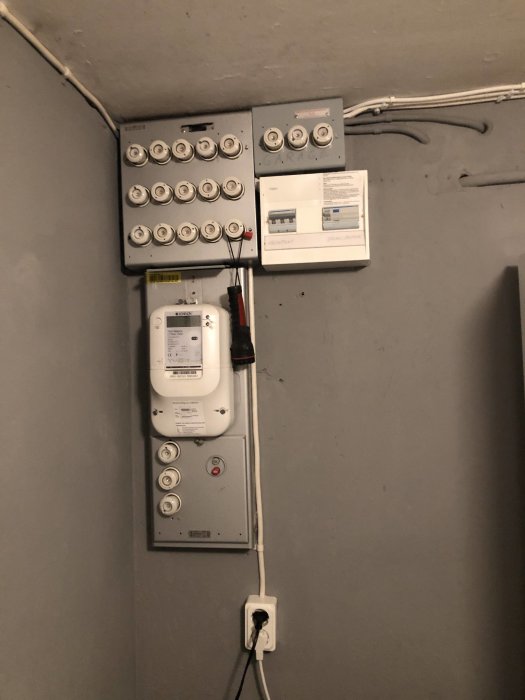 Ett proppskåp med säkringar och elektrisk mätare på en gråmålad vägg med kablar som leder uppåt och ut i rummet.
