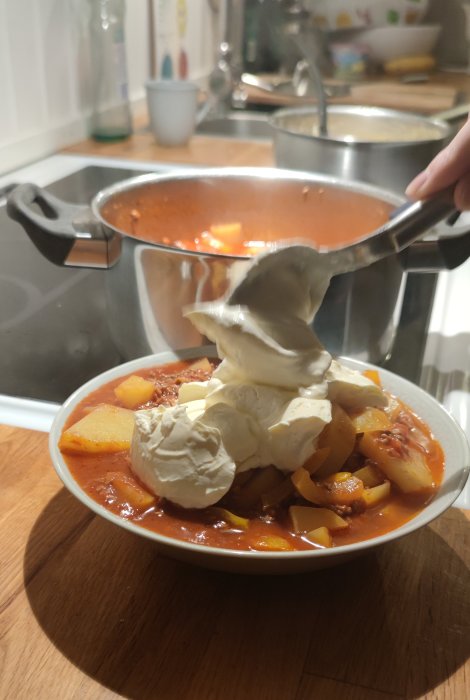 Portion av köttfärsgulasch med potatis och rikligt med creme fraiche på en tallrik, gryta i bakgrunden.