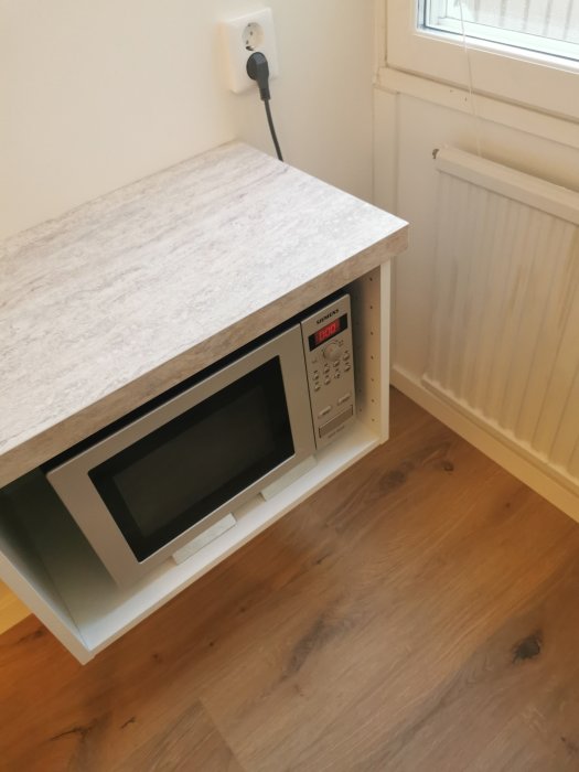 Mikrovågsugn kopplad till 10A köksuttag under bänkskiva, bredvid fönster och radiator.