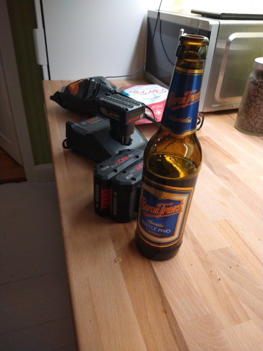En ölflaska bredvid en borr och batterier på ett träbord.