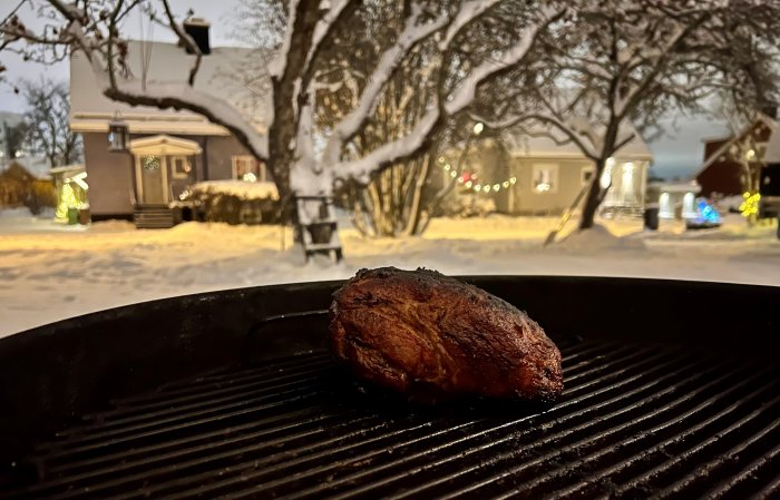 En stek grillas på en utomhusgrill med snötäckt trädgård och ljusslingor i bakgrunden.