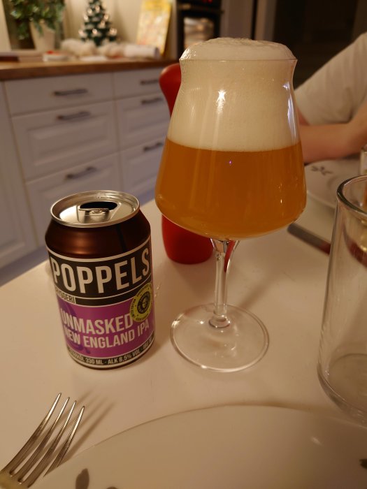 Ölglas med skummande ljus öl och en Poppels ölburk på ett vitt bord.