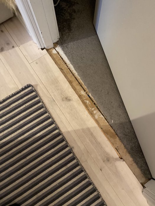 Dörröppning med synlig skadad golvyta där mattan möter trägolvet, tecken på tidigare renovering.