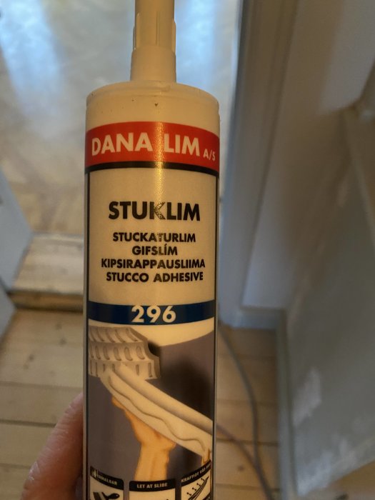 Hand håller en tub med DANA LIM stuklim för gips dekorationer, i en inomhusmiljö.