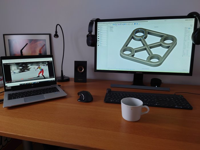 Två skärmar på ett skrivbord med CAD-program öppet och en video, tillsammans med en laptop, mus och kopp.