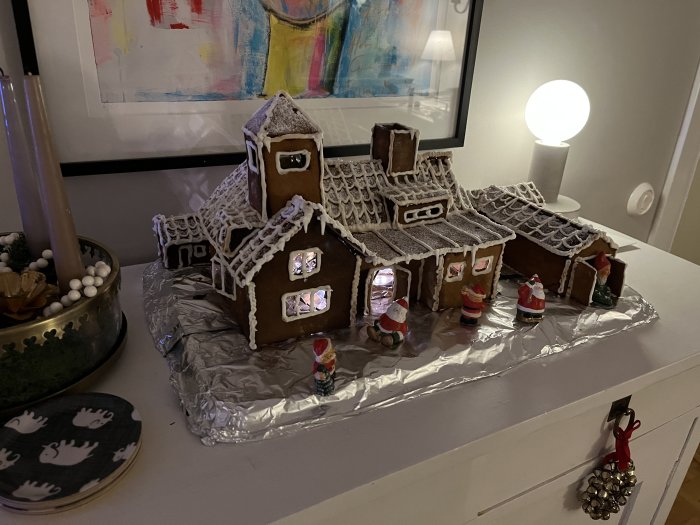 Handgjort pepparkakshus med dekorativ glasyr och små julfigurer framför på ett bord.
