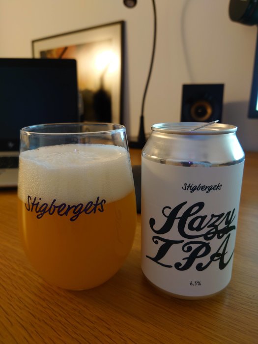 Ett glas fyllt med öl bredvid en burk märkt "Stigbergets Hazy IPA" på ett skrivbord.